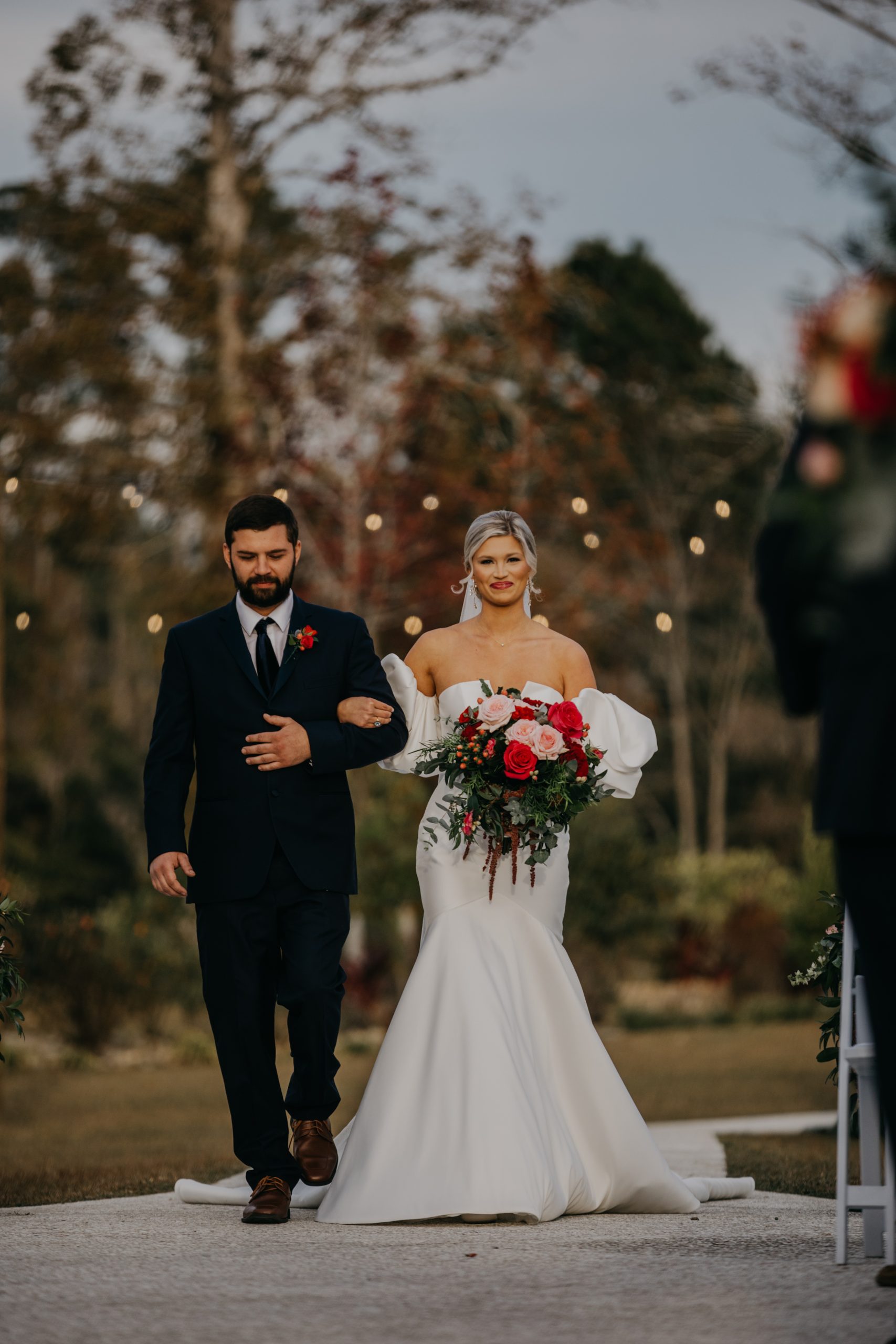 a bride walks down the aisle with her brother and she is carrying a huge red and pink wedding bouquet.