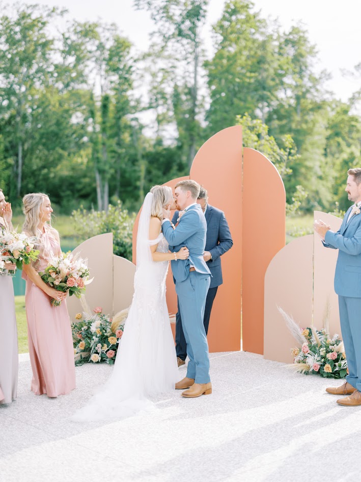 A trendy back drop for this bride and groom's first kiss at their wedding ceremony with bridesmaids dressed in pretty pink gowns holding lush boho style bouquets in coral hues.