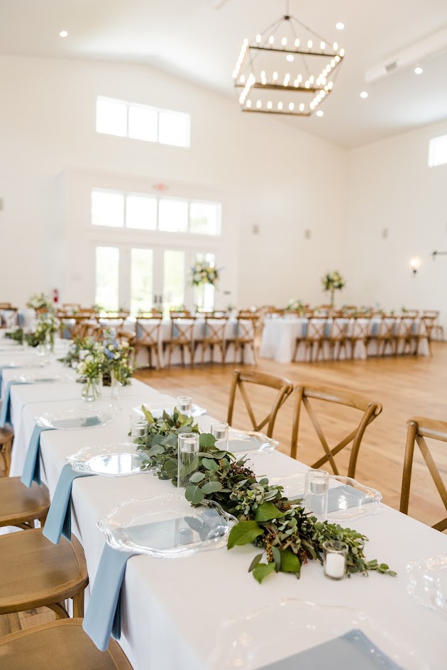 An elegant blue and white wedding table setup captures the perfect balance of timeless charm and modern sophistication. Crisp white linens set a clean, fresh foundation, while varying shades of blue in the wedding flowers and blue satin napkins. 