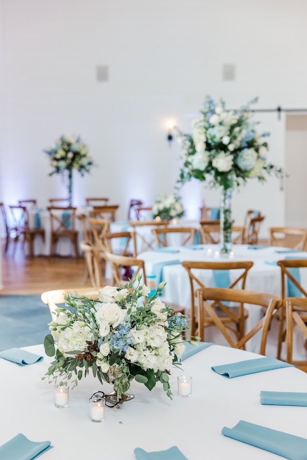Reception table set up with low lush florals in white and blue with greenery as an accent. Tall florals seen behind on additional tables. Dusty blue linens and votive candles make this elegant.