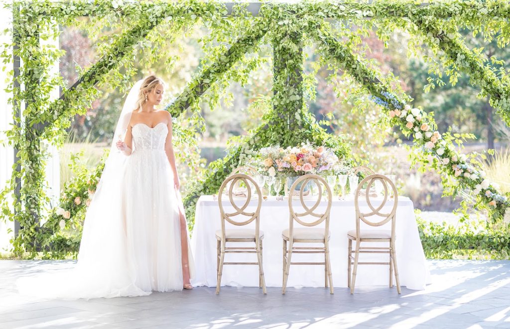 Bride in an elegant wedding gown standing beside a beautifully decorated table featuring pastel florals, fine china, and timeless stemware.