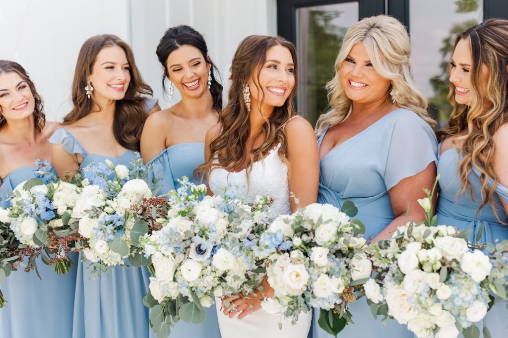 A bride in her gown and bridesmaids wearing pale dusty blue bridesmaid gowns holding their lush bridal bouquets in colors of blue and white smiling and looking at each other. 