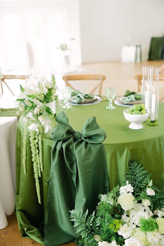 Sweetheart table decor in shades of green. A satin moss green linen covers the table with organic elements as decor and a swanky green satin bow showing on the side. Florals filled with whites and greens surround the table.