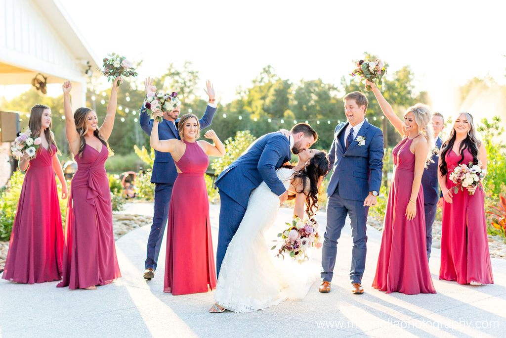 A bridal party wearing shades of mauve bridesmaid dresses along with their lush bouquets filled with white, blush , purple and pink florals cheering on the bride and groom as they kiss for their cheerful wedding portrait. 