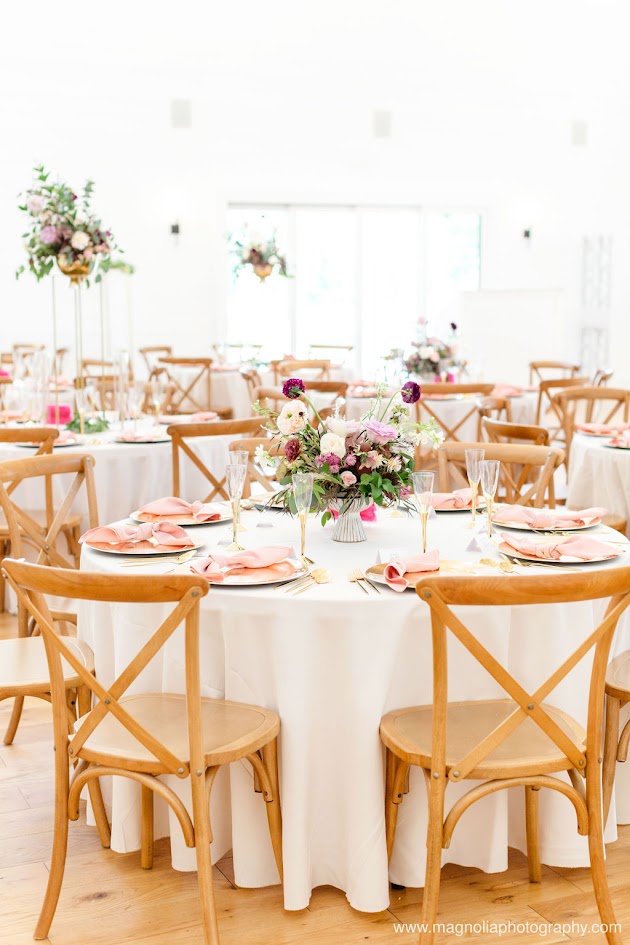 Pictured is an elegant wedding table setup where every detail feels romantic and refined. Soft shades of blush pink and delicate lavender create a graceful base, while accents of deep burgundy add richness and depth. Crisp white elements—whether in linens, florals, or fine china balance the palette, giving the tablescape a timeless sophistication. Layered textures like flowing fabrics, glowing candlelight, and carefully arranged blooms tie it all together, creating a design that feels both luxurious and inviting. This combination of pink, purple, burgundy, and white sets the stage for a celebration that’s as warm and romantic as it is unforgettable.